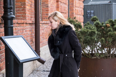 Young blonde woman in black winter coat with fur collar reads menu near a restaurant.