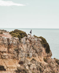 Distant view of man standing on cliff by sea against sky
