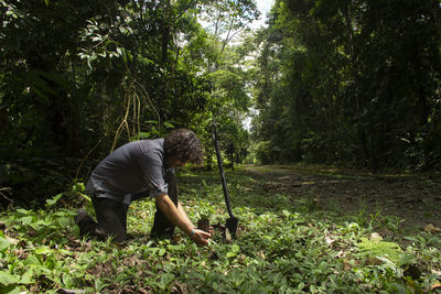 Hispanic man on his knees planting small plant with black shovel in green field surrounded by trees