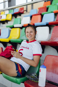 Happy player sitting on seat with smart phone at stadium