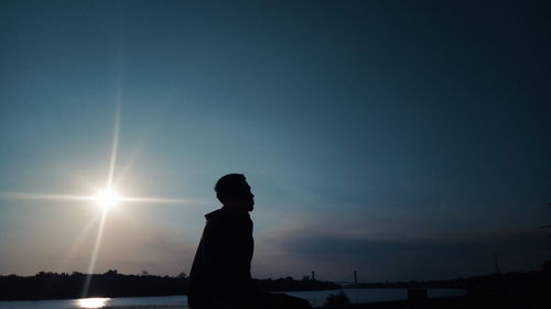 Side view of silhouette teenage boy sitting by lake against sky during sunset