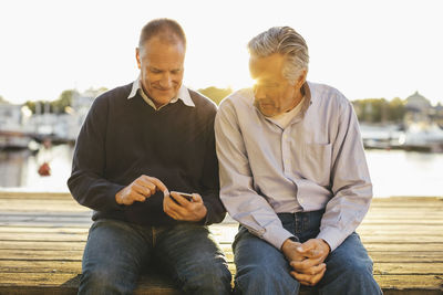 Senior man looking at male friend using mobile phone on pier