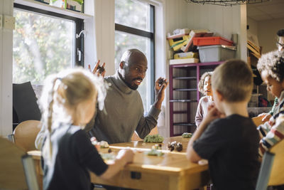 Male teacher explaining children while sitting at bench during clay molding class at kindergarten