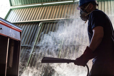 Low angle side view of male worker in hardhat and respirator using hose of fire extinguisher during accident in smoked workshop