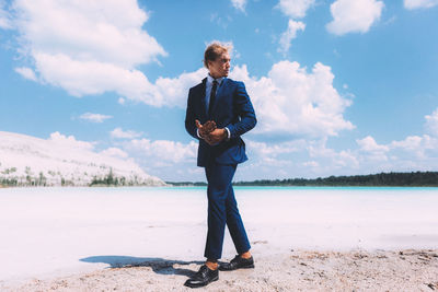 Full length of man with curly hair wearing suit while standing at beach against sky