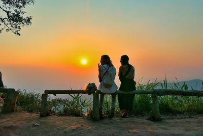 Female friends sitting on post against orange sky during sunset