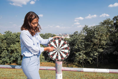 Young woman holding dart board against sky