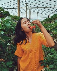 Woman eating strawberries while standing by plants in greenhouse