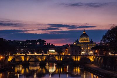 Illuminated bridge over river by buildings against sky at dusk