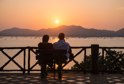 Rear view of couple sitting on shore against sky during sunset