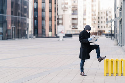 Full length of man reading newspaper while standing on footpath