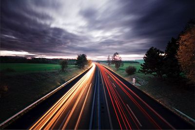 Light trails on road against cloudy sky