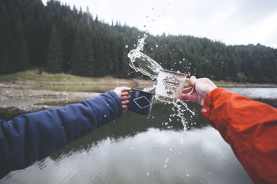 Cropped hands splashing water over lake against trees