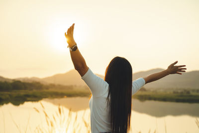 Rear view of woman with arms raised standing against sky during sunset