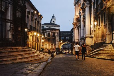 Rear view of man and woman walking on footpath amidst buildings at dusk