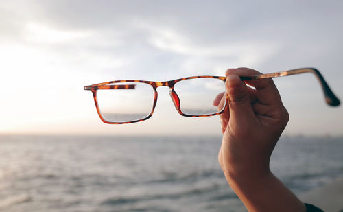Close-up of hand holding eyeglasses against sky at beach
