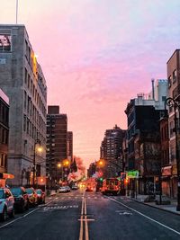 City street by buildings against sky during sunset