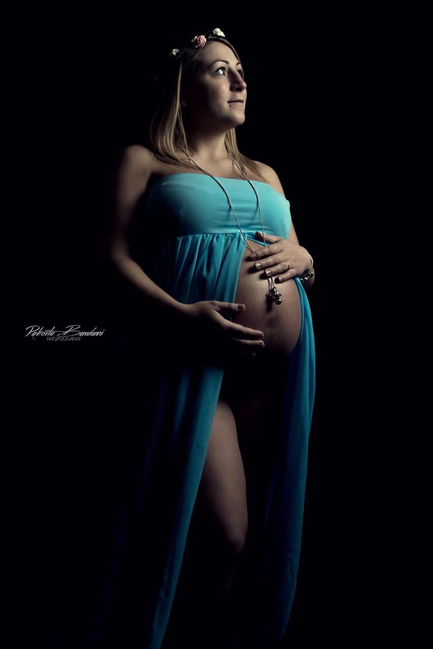 black background, studio shot, one person, young women, young adult, indoors, three quarter length, women, standing, human abdomen, beautiful woman, beauty, pregnant, clothing, adult, anticipation, side view, lifestyles, hairstyle, contemplation, human fertility