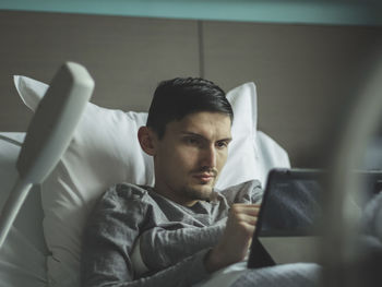 A young man with a tablet lies in bed in a hospital.