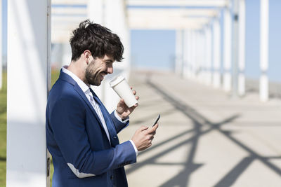 Happy young businessman with smartphone and takeaway coffee outdoors