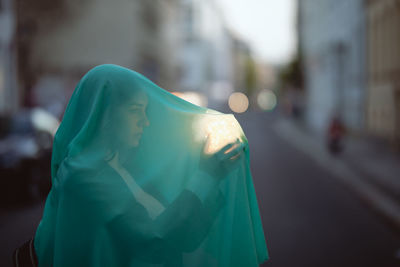 Woman holding glowing jar under blue scarf on city street at dusk