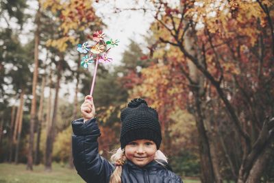 Portrait of smiling girl holding pinwheel against trees during autumn