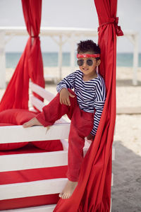 Stylish baby boy in a striped t-shirt and sunglasses stands at the red beach sunbed