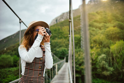 Woman holding camera while standing by tree