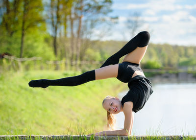 Side view portrait of smiling girl practicing handstand on retaining wall against lake during sunny day