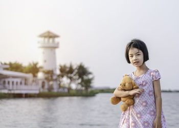 Girl holding teddy bear while standing against lake and sky