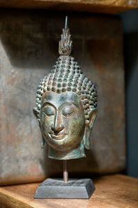 Brass buddha head mounted on a stand on a wooden shelf in the corner of a room with rustic walls