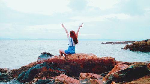 Woman with arms raised gesturing peace sign while sitting on rock by sea against sky