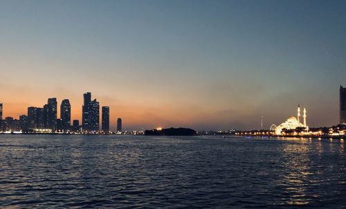 Sea by illuminated buildings against sky during sunset