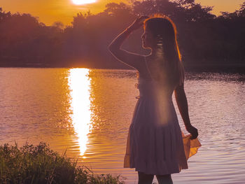 Rear view of woman standing against lake during sunset