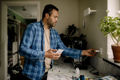 Man plugging smart phone charger in power socket while standing near kitchen counter at home