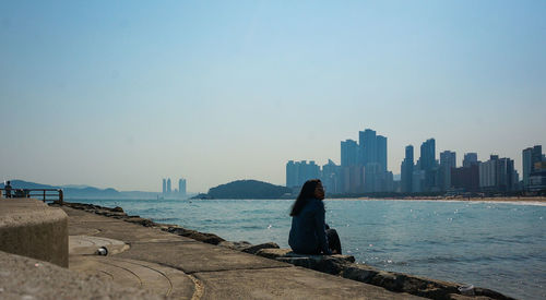 Woman sitting by sea in city against sky