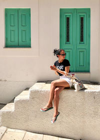 A young woman is sitting on white staircase leading to the white house with green door and shutters