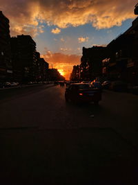 Cars on street amidst silhouette buildings against sky during sunset