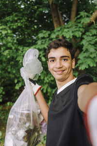 Portrait of smiling teenage boy showing garbage bag while taking selfie