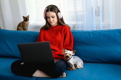 A young girl in a red  sweater works on a laptop at home. her cat and dog are sitting next to her