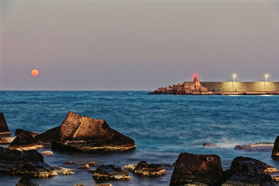 Rocks by sea against sky during sunset and moonrise