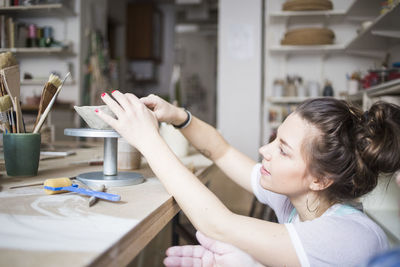 Side view of young female potter molding clay at workbench in store