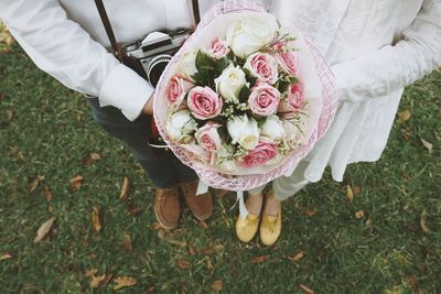 Low section of bride and groom holding bouquet while standing on grassy field