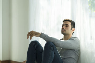 Young man looking away while sitting on window