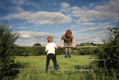 Full length of brothers on fence at grassy field against sky