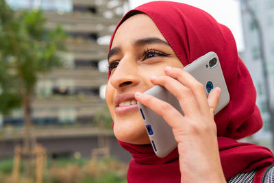 Low angle of positive ethnic female in hijab standing on street speaking on smartphone enjoying weekend