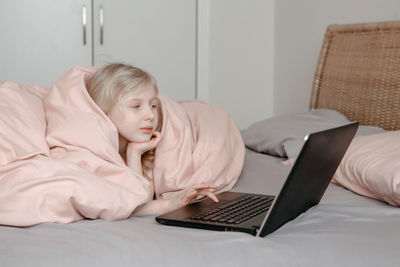 Bored girl lying in bed under blanket and learning in virtual online school class. 