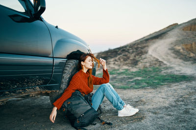 Young woman sitting on car against sky