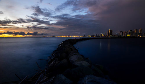 Sea by illuminated buildings against sky at sunset