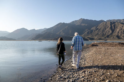 Senior man walking with his granddaughter along the lake shore with mountains.